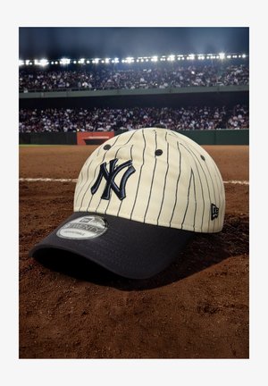 Beige Baseballkappe mit marineblauen Akzenten, vertikalen Nadelstreifen und einem gestickten Logo auf der Vorderseite. Verstellbare Größe.
