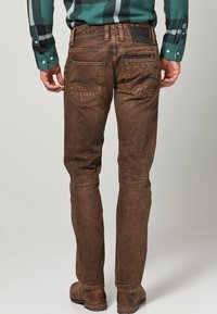 Jeans marrones de corte ajustado con textura desgastada, con bolsillos traseros y un detalle de cremallera en el lado derecho. Completados con un parche de logo negro.