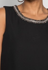 Haut noir sans manches avec un col orné de perles et embellissements, à la texture lisse. Le tissu est doux et léger.