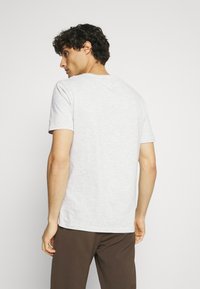Tricou din bumbac gri deschis, cu mâneci scurte, design clasic cu guler rotund, textură fină și spate simplu. Purtați cu pantaloni maronii.