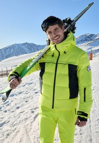 Felle gele ski-jas met zwarte accenten, voorzien van een capuchon, ritssluitingen en een logopatch. Draagt bijpassende skibroek in de sneeuw.