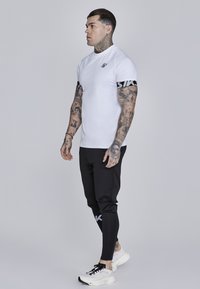 Hvid kortærmet skjorte med sorte accenter og logo, parret med sorte slim-fit bukser med branding, afsluttet med hvide sportsko.