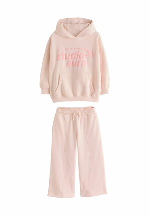 Young girl in pale pink hoodie and pants sitting cross-legged, smiling and looking up against a neutral background.