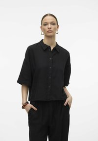 Camicetta nera a metà vita realizzata in un tessuto testurizzato, con chiusura a bottoni sul davanti, maniche corte e colletto classico. Il modello indossa pantaloni larghi.