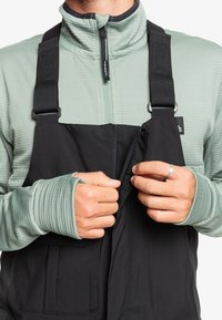 Pantalones de trabajo negros con tirantes ajustables, presentando una cremallera frontal y una camiseta de manga larga texturizada de color verde claro debajo.