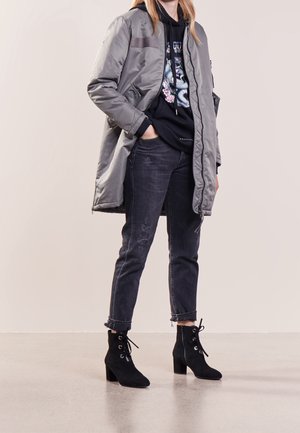 Femme portant une longue veste grise par-dessus un sweat à capuche noir avec un motif serpent, un jean sombre roulé aux chevilles et des bottines noires à talons.