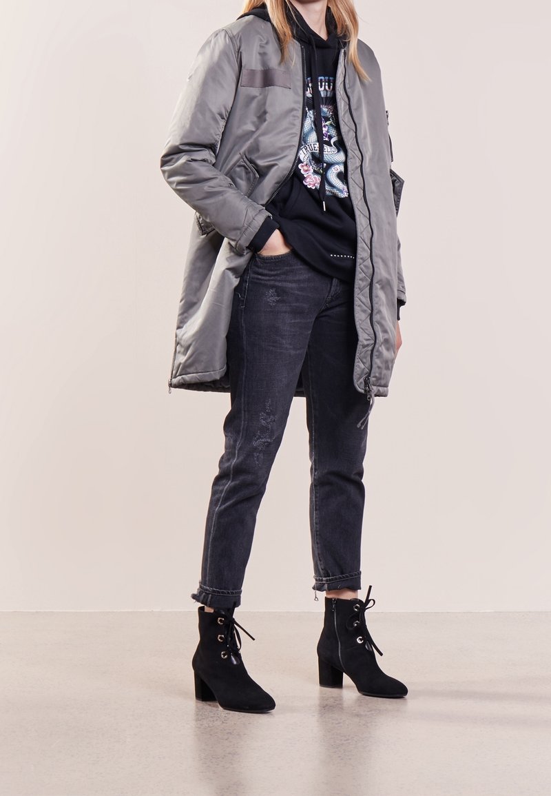 Femme portant une longue veste grise par-dessus un sweat à capuche noir avec un motif serpent, un jean sombre roulé aux chevilles et des bottines noires à talons.