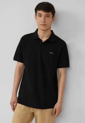 Schwarzes Poloshirt mit kurzen Ärmeln und klassischem Kragen, versehen mit einem kleinen goldenen Logo auf der linken Brust, präsentiert mit khakifarbener Hose.