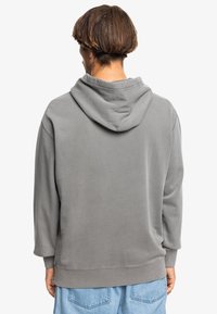 Felpa grigia con un cappuccio ampio, polsini e fondo a costine. Tessuto liscio, vestibilità casual, indossata con denim azzurro chiaro.