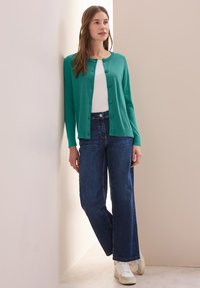 Türkiser Strickjacke mit Knöpfen, kombiniert mit hoch geschnittenen, dunklen Denim-Jeans und weißen Sneakers, vor einer neutralen Wand stehend.