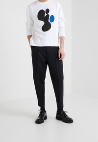 Homme portant un sweat-shirt blanc avec des formes abstraites noires et bleues, un pantalon noir court, des chaussettes noires et des chaussures en cuir noires à lacets.