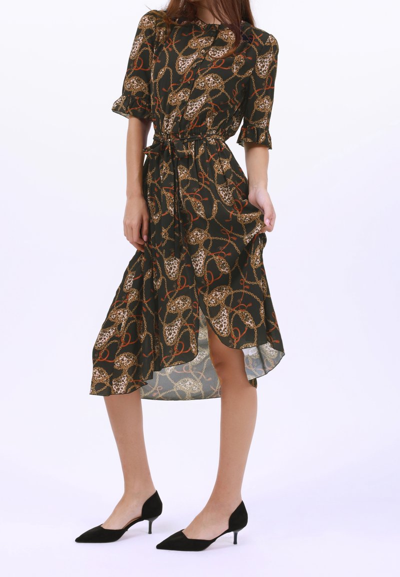 Robe vert foncé avec une chaîne et un imprimé léopard, manches trois-quarts, taille ceinturée, et ourlet asymétrique. Associée à des talons noirs.