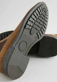 Zapato de ante marrón que muestra una suela de goma negra texturizada con un patrón de cinta. La suela presenta un diseño circular en la sección media.