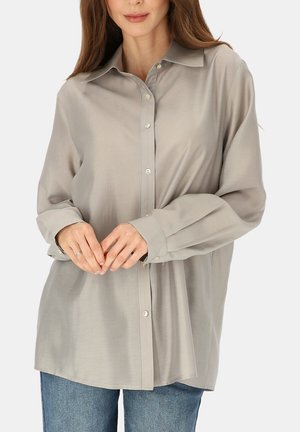 Kobieta ubrana w luźną, jasnoszarą koszulę na guziki z długimi rękawami i niebieskie dżinsy, ręce delikatnie splecione na talii.