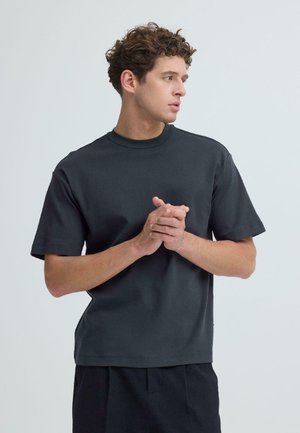 Ung mand med krøllet hår iført sort t-shirt og mørke bukser, der holder hænderne sammen og kigger til siden mod en ensfarvet baggrund.