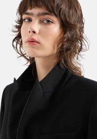 Cappotto nero con colletto alto e rever angolari, caratterizzato da una trama liscia e una vestibilità sartoriale, indossato sopra un top semplice.