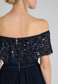 Námořnické off-shoulder šaty s texturovaným krajkovým korpusem zdobeným flitry a květinovými vzory, s viditelným zipem na zadní straně.