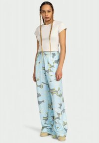 Pantaloni a gamba larga di colore azzurro chiaro con un motivo floreale in tonalità grigio e verde, abbinati a una maglietta attillata bianca a maniche corte.