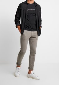 Man i svart skjorta över en svart Tommy-t-shirt, taupe slim-fit byxor uppvikta vid ankelna, och vita sneakers som står mot en vit vägg.