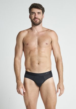 Skægget mand med kort mørkt hår iført sorte underbukser, stående mod en ensfarvet lys grå baggrund, vendt fremad med et neutralt udtryk.