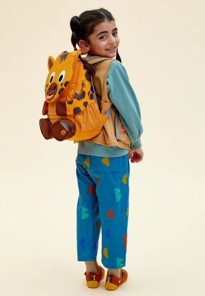 Smiling child with pigtails wearing colorful pants, a teal sweatshirt, a tan vest, red shoes, and an orange animal-themed backpack.