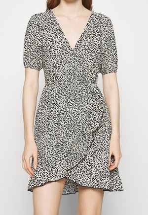 Femme portant une robe portefeuille courte à imprimé léopard noir et blanc, manches bouffantes et ourlet à volants, debout devant un fond uni.