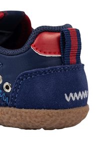 Zapato azul marino con parte superior de materiales mixtos, acentos rojos y una suela de goma texturada marrón. Presenta costuras y ojales blancos en contraste.