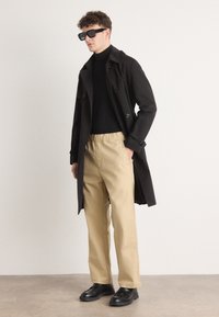 Manteau trench noir à double boutonnage, col roulé noir ajusté, pantalon beige à taille élastique et chaussures en cuir noires. Lunettes de soleil mate.