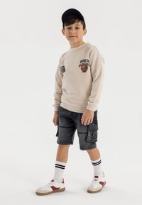 Beige sweatshirt med grafiken "Brooklyn Basketball", grå cargo-shorts, vita och röda sneakers samt svart keps. Vita strumpor med svarta ränder.