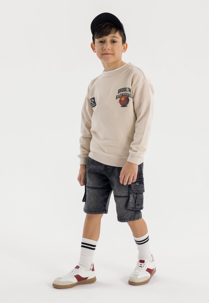Beige sweatshirt med grafiken "Brooklyn Basketball", grå cargo-shorts, vita och röda sneakers samt svart keps. Vita strumpor med svarta ränder.