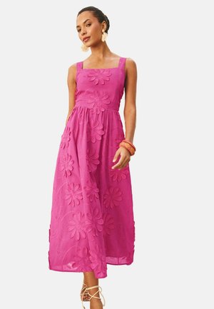 Vrouw die een mouwloze roze maxi-jurk draagt met grote bloemenborduurwerk, staand met één ontspannen hand en bladvormige oorbellen.