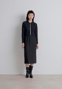 Schwarzes Midikleid mit langen Ärmeln, Reißverschluss und plissierter Textur. Getragen mit knielangen schwarzen Stiefeln. Glatter Stoff mit taillierter Passform.