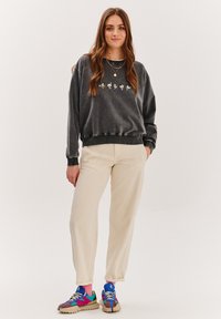 Šedá mikina s grafickým designem, béžové kalhoty a vícero barevné tenisky. Materiál vypadá měkce, s volným střihem a žebrovanými manžetami.