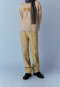 Pull beige avec motif de palmier, assorti à un pantalon clair et ample. Complété par des chaussures noires et une écharpe rayée foncée.