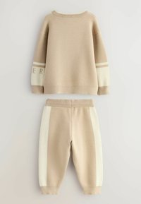 Beige gebreide trui en bijpassende joggingsbroek. De trui heeft geribbelde manchetten en een streep op de mouwen; de broek heeft zijkant kleurblokken.