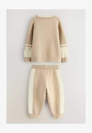 Beige kudumüts ja sobivad jogger-püksid. Kudumütsil on soonikkoes varrukad ja triip varrukatel; püksid on külgpaneelide värvilised.