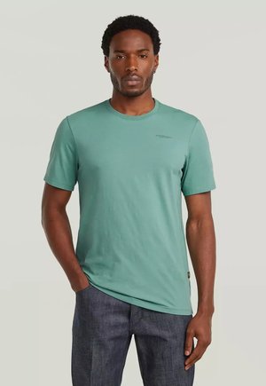 Uomo con t-shirt turchese a girocollo e jeans blu scuro, in piedi con la mano sinistra in tasca davanti a uno sfondo semplice.