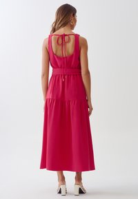 Pink sleeveless maxi dress with a tied open back, cinched waist, and gathered skirt. Features a smooth texture and subtle sheen.