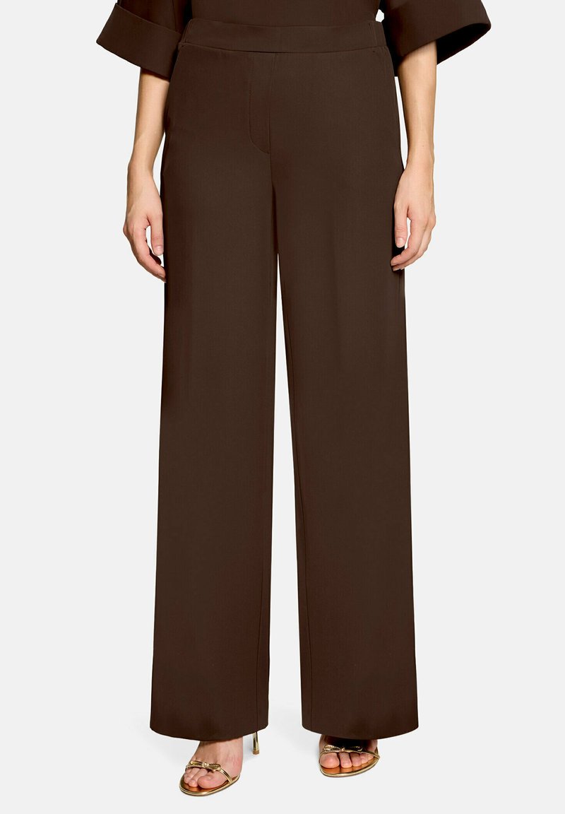 Femme portant un pantalon marron foncé taille haute à jambes larges et des sandales ouvertes dorées à talons, debout sur fond blanc.