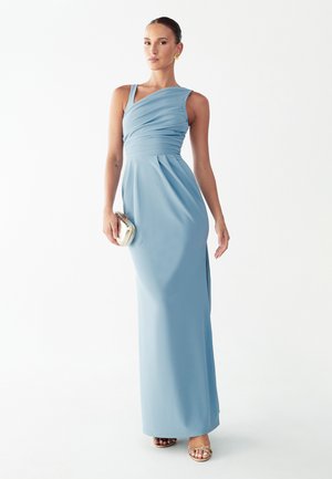 Frau in einem bodenlangen, hellblauen Ein-Schulter-Kleid, die eine kleine goldene Clutch hält und goldene Sandalen sowie goldene Ohrringe trägt.