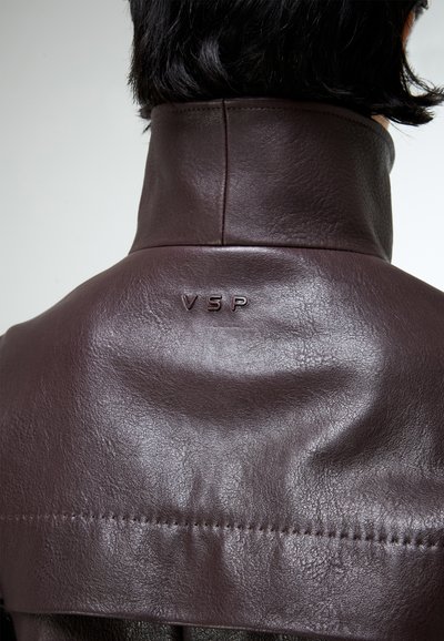 Rövid fekete hajú személy sötétbarna bőrkabátot visel, amelynek felhajtott gallérja és a hátán dombornyomott "VSP" betűk találhatók.