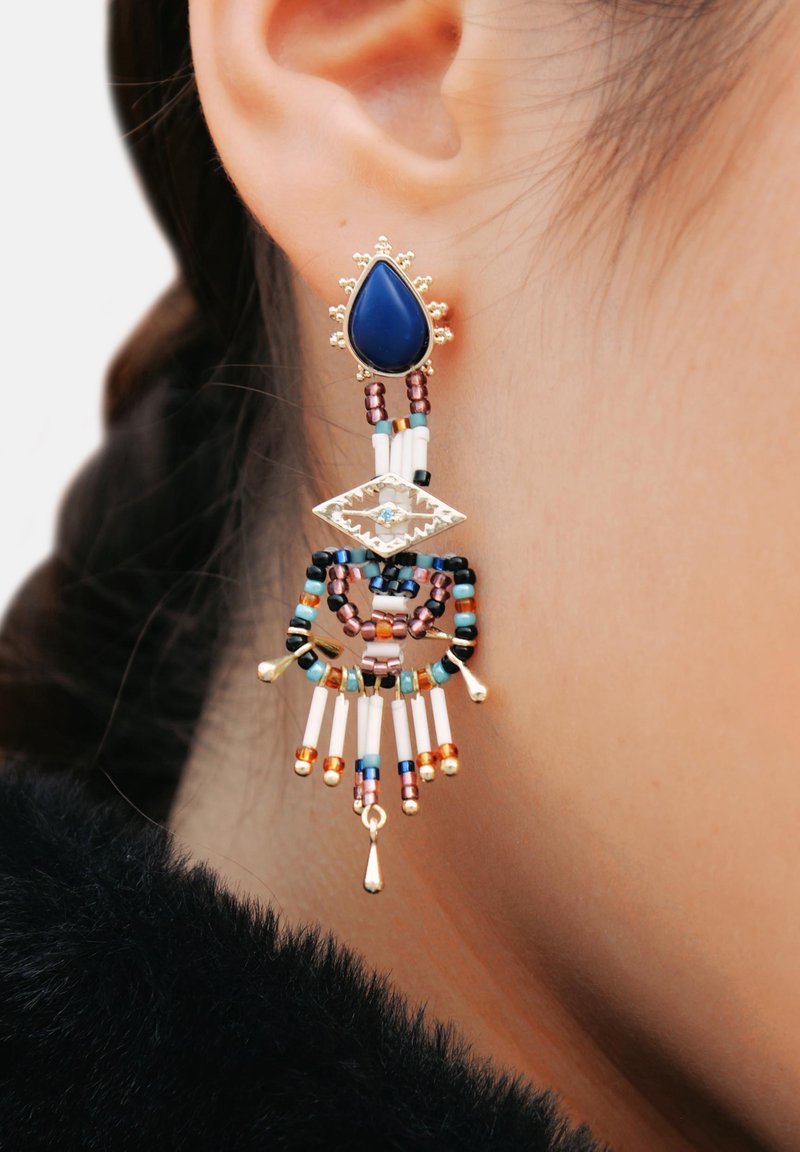 Boucles d'oreilles dorées avec une pierre en forme de goutte d'eau bleu, un travail de perles complexe dans différentes couleurs, et des accents pendants avec un design découpé.