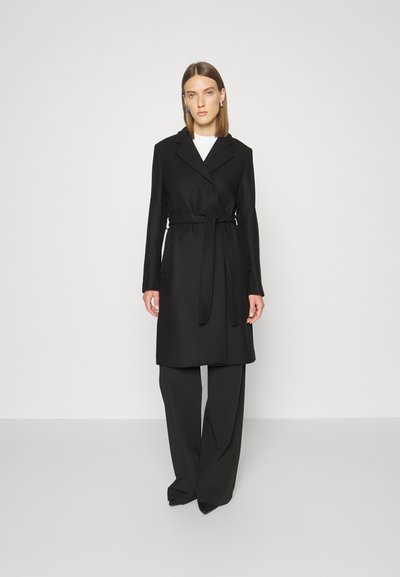 Black wrap coat in a tailored design with a belt at the waist, featuring long sleeves and a notched collar, paired with black wide-leg trousers.