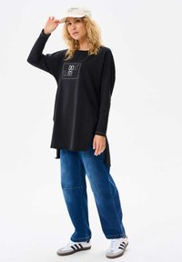 Czarna koszula z długim rękawem z napisem "NIE PODDAWAJ SIĘ!"; połączona z luźnymi niebieskimi dżinsami i białymi trampkami w szare paski. Kremowa czapka dopełnia całość.