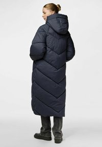 Dlouhá námořnická péřová bunda s kapucí, s šípovým prošíváním a volným střihem. Nositá přes šedé džíny a tmavé boty.