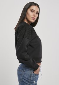 Felpa nera corta con spalle in pizzo e polsini elasticizzati, abbinata a jeans blu strappati. Design semplice e linee pulite.
