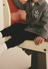 Enfant portant une veste grise avec les lettres "LA" assis sur un banc rouge et blanc, jambes étendues, mains reposant à côté.