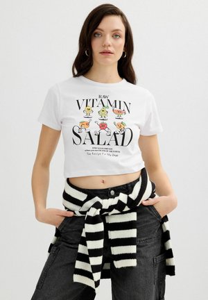 Joven mujer con un crop top blanco con frutas de dibujos animados coloridas y el texto "Raw Vitamin Salad", pantalones negros y un suéter de rayas blanco y negro atado a la cintura.