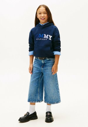 Fille souriante, portant un sweat à capuche marine avec l'inscription "Tommy Hilfiger", un jean large bleu, des chaussettes blanches et des mocassins noirs, debout devant un fond uni.