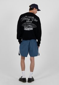Mann mit einem schwarzen Pullover mit einem Racing Club Berlin Auto- und Globus-Design, blauen Shorts, weißen Socken, schwarzen Schuhen und einer marineblauen Kappe, der nach hinten blickt.